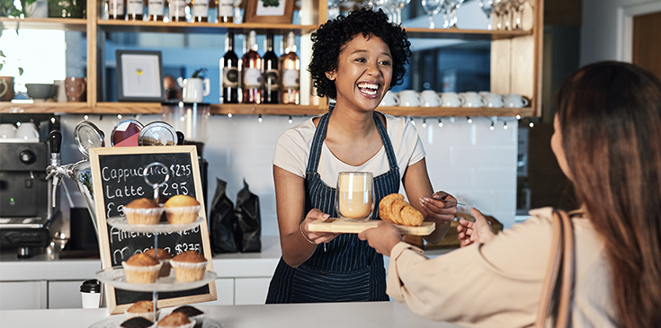 Barista hinter dem Tresen eines Cafés reicht einer Kundin ein Tablett mit Kaffee und Croissant.