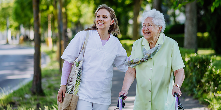 Eine ältere Frau mit Rollator geht lächelnd an der Seite einer jüngeren Begleiterin auf einem sonnigen, von Bäumen gesäumten Gehweg.