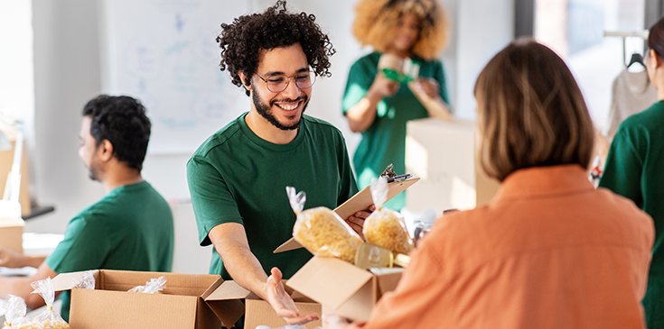 Fröhlich lächelnder männlicher Helfer mit Klemmbrett und eine Frau, die bei einer Lebensmittelverteilung oder Flüchtlingshilfe eine Kiste mit Lebensmitteln entgegennimmt.
