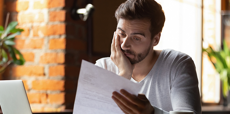 Ein Mann sitzt an einem Tisch in einem Café oder Büro, hält ein Blatt Papier in der Hand und schaut mit nachdenklichem, leicht besorgtem Ausdruck darauf.