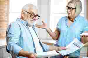 Ein älterer Mann mit Brille blickt verwundert auf Papierunterlagen, die er in seinen Händen hält. Seitlich rechts neben ihm steht eine ältere Frau, auch mit Brille, die ihre Arme verwundert hochzieht und den Mann dabei anschaut.