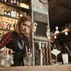 Eine Frau im karierten Hemd steht hinter einer Baar und mixt Cocktails. Sie hat einen kleinen Haardutt und trägt eine Schürze. Im Hintergrund sind verschwommen Getränkeflaschen, Gläser und Lichte zu erkennen.