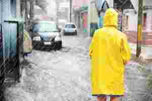 Person von hinten in gelbem Regenmantel steht in strömendem Regen auf einer überschwemmten Straße und schaut auf Häuser und Autos.