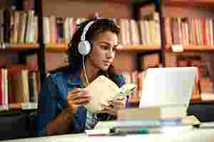 Eine junge Frau mit braunen, lockigen Haaren sitzt in der Bibliothek an einem Schreibtisch. Sie schaut auf den aufgeklappten Laptop und trägt Kopfhörer. In ihrer Hand hält sie ein Buch, auch um sie herum sind Bücher zu erkennen.