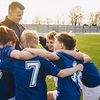 Ein Trainer und sein Team aus Jungs stehen im Kreis und haben die Arme über die Schultern der anderen gelegt. Alle lächeln.