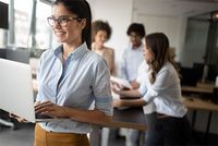 Aktuelles BFH-Urteil: Kindergeld in der Erst- und Zweitausbildung Eine junge Frau in Bluse und Hose träft eine Brille. Auf dem Arm hat sie einen Laptop und sie schaut schräg aus dem Bild heraus. Sie lächelt aufgeregt. Im Hintergrund sind verschwommen drei weitere Personen zu sehe, die an einem Projekt arbeiten.