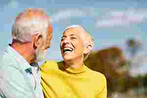 Eine ältere Frau mit kurzen Haaren und in Rollkragenpulli und ein älterer Mann mit Hemd umarmen sich. Der Mann ist eher von hinten zu erkennen; die Frau lächelt über das ganze Gesicht. Im Hintergrund befinden sich Bäume, die verschwommen sind.
