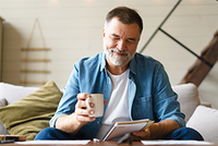Lächelnder Mann mittleren Alters sitzt mit einer Tasse Kaffee auf dem Sofa und schaut auf sein Smartphone.