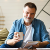 Lächelnder Mann mittleren Alters sitzt mit einer Tasse Kaffee auf dem Sofa und schaut auf sein Smartphone.