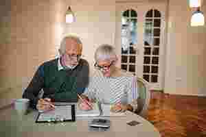 Ein älterer Mann und eine älteren Frau sitzen in ihrer Wohnung an einem Tisch und füllen Formulare aus. Auf dem Tisch befindet sich noch eine Tasse und ein Taschenrechner.