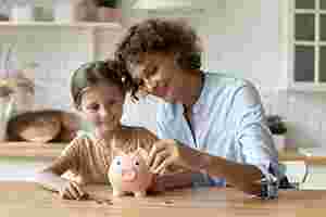 Eine Mutter und ihre kleine Tochter werfen lächelnd Geldmünzen in ein rosafarbenes Sparschwein.