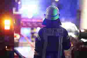 Ein Mann mit Helm und Feuerwehruniform ist von hinten zu sehen. Links neben ihm und verschwommen im Hintergrund sind Feuerwehrautos zu erkennen.
