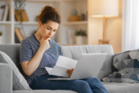 Eine junge Frau prüft ihren Steuerbescheid. Sie sitzt auf dem Sofa und hat ihren Laptop auf ihren Beinen.