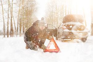 Ein Mann mit Mütze, Handschuhen und dicker Jacke kniet auf einer schneebedeckten Straße und stellt ein Warndreieck auf. Hinter ihm steht sein Auto mit geöffneter Tür und Kofferraum. Es fällt viel Schnee.