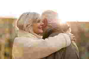 Eine lächelnde, ältere Frau in Wollmantel und Schal umarmt einen älteren Mann. Dessen Gesicht ist nicht zu erkennen. Der Hintergrund ist verschwommen.