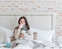 Husten, Schnupfen, Heiserkeit: Fragen Sie Ihren Arzt oder Steuerberater Eine unglückliche, kranke Frau sitzt mit Schal im Bett.