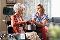 Pflegekosten als außergewöhnliche Belastungen absetzen Eine Frau sitzt auf dem Sofa und hält die Hand ihrer, im Rollstuhl sitzenden, Mutter. Beide Frauen lächeln sich gegenseitig an.