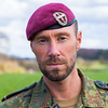 Soldat in Tarnuniform mit bordeauxrotem Barett blickt in die Kamera, im Hintergrund eine grüne Landschaft.