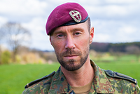Soldat in Tarnuniform mit bordeauxrotem Barett blickt in die Kamera, im Hintergrund eine grüne Landschaft.