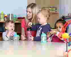 Kinderbetreuung: Junge Erzieherin beschäftigt sich mit drei kleinen Kindern am Tisch.