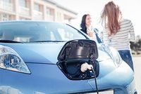Neue Steuerregeln für dienstliche Elektroautos Im Vordergrund des Bildes befindet sich ein Elektroauto, das gerade aufgeladen wird. Neben dem Auto stehen leicht verschwommen zwei lächelnde Frauen, von denen eine nur von hinten zu erkennen ist.