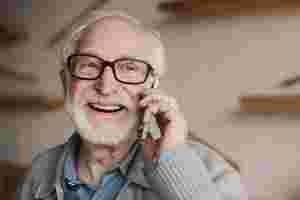 Ein älterer Mann mit Bart, Brille und beiger Strickjacke telefoniert angeregt mit seinem Handy. Im Hintergrund sieht man unscharf die Umrisse seiner Wohnung. 