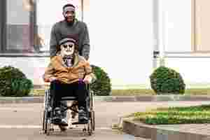 Ein Mann in Pulli schiebt einen älteren Mann mit Jacke und Sonnenbrille in einem Rollstuhl über die Straße. Beide lächeln und sind gut gelaunt.