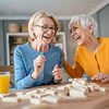 Zwei ältere Frauen lachen gemeinsam beim Spielen eines Gesellschaftsspiels an einem Tisch.