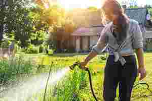 Eine junge Frau bewässert ihre Pflanzen mit einem Gartenschlauch, im Hintergrund steht ein Gartenhaus.