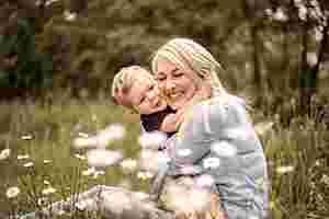 Eine blonde Frau sitzt auf einer Blumenwiese und umarmt liebevoll ihren kleinen Sohn. Der Sohn hat seine Arme um den Hals seiner Mutter gelegt. Beide lächeln.