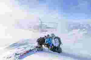 Drei Personen in Ski-Montur sitzen auf einem schneebedeckten Berg und schauen auf eine Karte. Im Intergrund sind weitere schneebedeckte Berge, blauer Himmel und ein Helikopter zu sehen, der Schnee aufwirbelt.