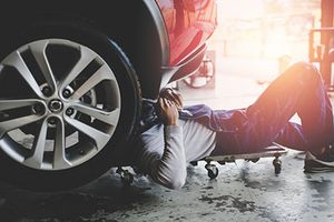 In einer Autowerkstatt fährt ein Kfz-Mechaniker auf dem Rücken auf einem Rollbrett unter ein Auto, dessen rechter Vorderreifen großzügig auf dem Bild platziert ist. Der Kopf des Arbeiters verschwindet unter dem Auto.