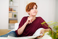 Junge Person mit kurzen roten Haaren sitzt entspannt auf einem Sofa und hält nachdenklich einen roten Stift ans Kinn.