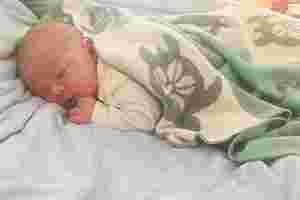 Ein neu geborenes Baby liegt in einer Decke eingewickelt auf einem Bettlaken. Das Baby hat die Augen geschlossen, die Hand an seinem Kinn und schläft friedlich.