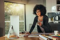 Corona-Virus: Home-Office von der Steuer absetzen Eine Frau befindet steht an einem großen Tisch und arbeitet. Auf dem Tisch sind ein großer PC, Unterlagen und Stifte platziert. Im Hintergrund ist eine Küche zu erkennen, sowie Stühle und ein großes Fenster.