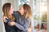 Unterhaltszahlungen an erwachsene Kinder in der Ausbildung Eine junge Frau und eine ältere Frau umarmen sich. Sie tragen Strickklamotten und lachen sich an. Im Hintergrund erkennt man verschwommen Pflanzen und Fenster.