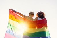 Zwei Frauen sind von hinten zu sehen. Sie haben ein großes Tuch in Regebogen-Darben um sich gelegt und blicken der Sonne entgegen.