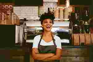 Eine Frau in T-Shirt und Schürze steht vor einer Kaffeetheke. Sie lacht, hat die Arme verschränkt und scheint eine Angestelltes des Cafés zu sein.