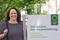 Kein Tag wie der andere: Interview mit Melanie Früchtenicht, Steuerring-Bereichsleiterin Eine Frau in gemusterter Bluse steht neben einem Schild, beschriftet mit Steuerring-Logo und „Wir machen Ihre Steuererklärung“.