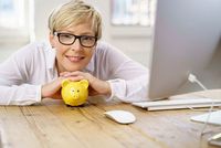 Mit vielen Versicherungen Steuern sparen Frau mit Brille und blonden, kurzen Haaren sitzt an einem Holztisch und stützt sich auf ein gelbes Sparschwein