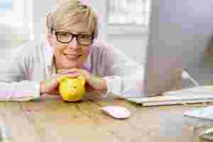 Frau mit Brille und blonden, kurzen Haaren sitzt an einem Holztisch und stützt sich auf ein gelbes Sparschwein