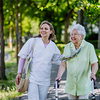 Eine ältere Frau mit Rollator geht lächelnd an der Seite einer jüngeren Begleiterin auf einem sonnigen, von Bäumen gesäumten Gehweg.