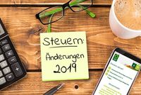 Steuern 2019: Was ändert sich? Ein gelber Notizzettel klebt auf einem Holztisch, darauf steht „Steuern: Änderungen 2019“. Um den Notizzettel herum liegen ein Taschenrechner, ein Kugelschreiber, eine Lesebrille und ein Smartphone mit der geöffneten Webseite des Steuerrings. Eine Tasse Milchkaffee rundet das Bild ab.