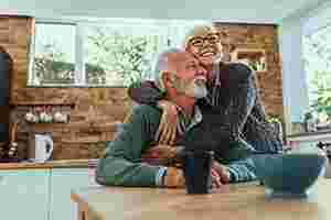 Ein älterer Mann sitzt am Küchentisch, eine ältere Frau mit Brille steht hinter ihm und hat ihre Arme um seine Schultern gelegt. Sie freuen sich und lächeln. Im Hintergrund befinden sich eine Küchenzeile und ein Fenster, durch das Bäume zu erkennen sind.