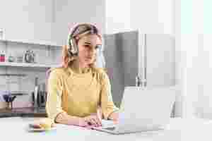 Eine Frau sitzt am Küchentisch und arbeitet am Laptop. Sie hat Kopfhörer auf und neben ihr steht ein kleiner Teller mit Snacks. Im Hintergrund befindet sich ihre Küche, die leicht verschwommen ist.