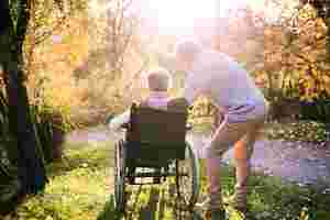 Eine Frau sitzt im Rollstuhl, der am Waldrand steht. Hinter ihr steht ihr Mann und bückt sich zu ihr herunter.