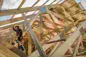 Ein Bauarbeiter mit Helm und Werkzeug steht auf einem Holzdach, das gerade ausgebaut wird. Durch das Dach hindurch sieht man den blauen Himmel