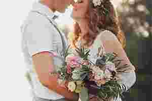 In der rechten Bildhälfte steht ein Hochzeitspaar, das sich anschaut und seine Gesichter aneinanderschmiegt. Die Braut trägt die Haare gewellt offen und hat Blumen im Haar, in der Hand hält einen Blumenstrauß. Der Bräutigam hat seine Hand auf die Hüfte der Braut gelegt.