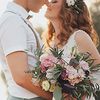 In der rechten Bildhälfte steht ein Hochzeitspaar, das sich anschaut und seine Gesichter aneinanderschmiegt. Die Braut trägt die Haare gewellt offen und hat Blumen im Haar, in der Hand hält einen Blumenstrauß. Der Bräutigam hat seine Hand auf die Hüfte der Braut gelegt.