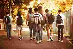 Eine Gruppe von Jugendlichen läuft nebeneinander eine Straße entlang. Sie tragen Rucksäcke. Im Hintergrund sind Bäume zu sehen.